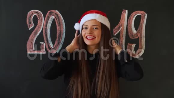 年轻女子在圣诞老人的帽子上的石板墙背景与铭文2019年女孩微笑着她会很高兴视频的预览图