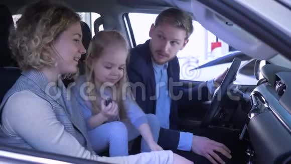 家庭在汽车销售中心快乐的微笑爸爸妈妈和小女孩回顾汽车和挥动钥匙坐着视频的预览图