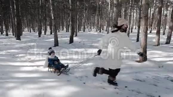 一位年轻活跃的母亲在森林中奔跑拉着雪橇她的孩子坐在雪橇上视频的预览图