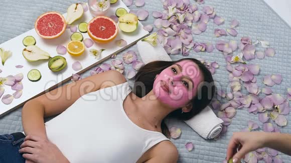 美丽的女人脸上戴着粉红色的面具而女性美容师在健康中心把黄瓜放在眼睛上视频的预览图