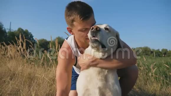 年轻人在草地上拥抱和抚摸他的拉布拉多他在大自然中和他的宠物在一起爱情和友谊视频的预览图
