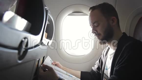 一个没有刮胡子的人正在舷窗附近的飞机上看杂志视频的预览图