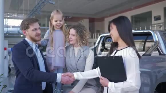 成功的汽车销售幸福的女经理与汽车陈列室的家庭买家握手微笑的表情视频的预览图