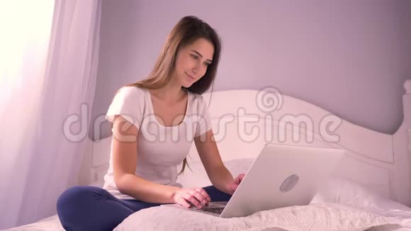 年轻迷人的少女坐在床上微笑着在笔记本电脑上打字的特写照片然后看着视频的预览图
