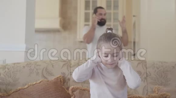 肖像小女孩坐在沙发上捂着耳朵因为她爸爸在电话里大声说话视频的预览图