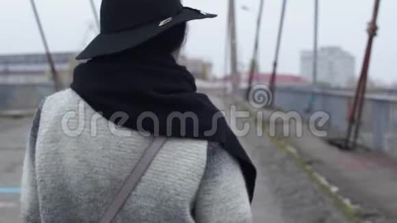 戴着宽边帽子的成年妇女在寒冷的多风灰色天气中穿过大桥转身视频的预览图