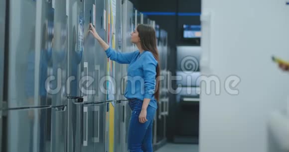 年轻性感的黑发女人穿着衬衫打开电器商店冰箱的门并与其他人进行比较视频的预览图