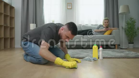 中镜头男人清洁地板女人躺在沙发上用智能手机视频的预览图