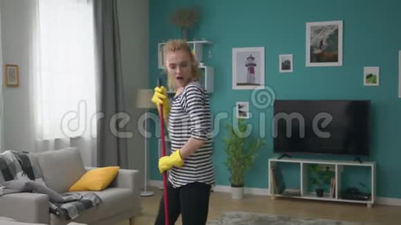 年轻女子在公寓里洗地板像麦克风一样用拖把唱一首歌视频的预览图