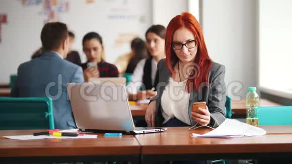 一个戴眼镜的姜女坐在办公室在笔记本电脑旁工作检查她的手机然后把它移开视频的预览图
