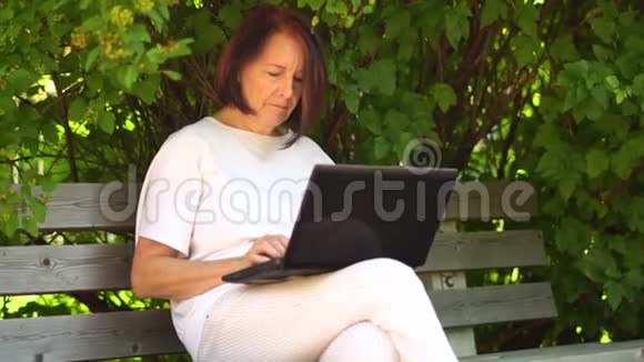 中年妇女在户外的笔记本电脑上工作微笑的中年祖母在电脑上遥远地工作视频的预览图