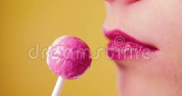 一个嘴唇粉红色的女人甜美地舔一个棒棒糖喜欢吃糖果黄色背景微笑有趣工作室视频的预览图