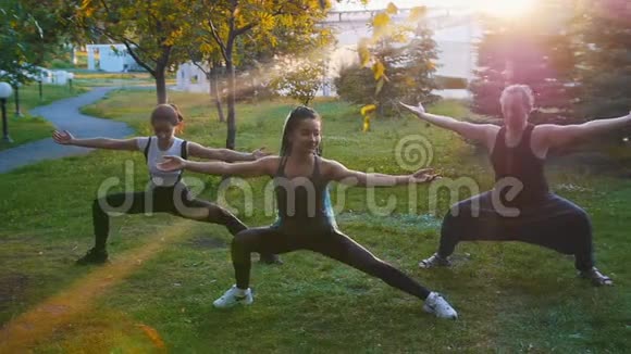 两位年轻女性在阳光下和教练一起在公园里做瑜伽体式一位女性有着长长的蓝色恐惧症视频的预览图
