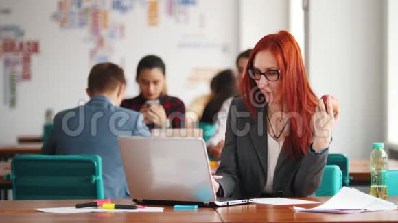 一个戴眼镜的姜女坐在办公室在笔记本电脑旁工作吃苹果视频的预览图