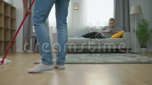 疲惫的男人拖地板女友在沙发上玩智能手机视频的预览图