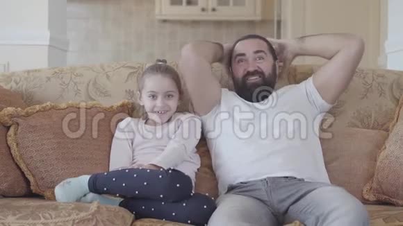 可爱的小女儿和她有趣的父亲一起在电视上观看有趣的电影在舒适的客厅里带着巨大的情感视频的预览图