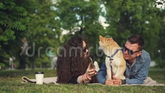 年轻女子带着宠物狗在公园放松时在智能手机上给男友展示有趣的照片人们都是视频的预览图