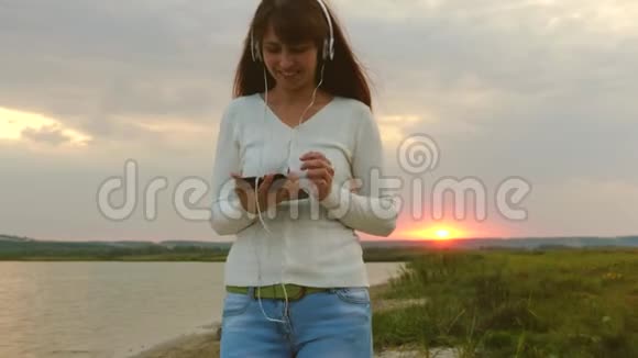 快乐女孩在平板电脑上看视频在湖边公园散步戴着耳机的漂亮女孩在沙滩上散步视频的预览图