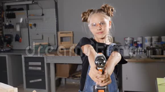 可爱的女孩穿着围裙拿着木工钻一个拿着钻头看镜头的小女孩的肖像视频的预览图