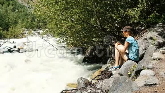 女人坐在一块石头上看着一条沿着森林流淌的山间河流缓慢的运动视频的预览图