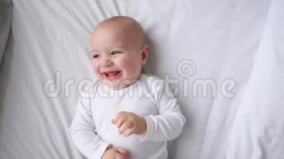 一个穿着白色t恤的孩子躺在一张白色的床上看着摄像机慢镜头地笑着俯视着视频的预览图