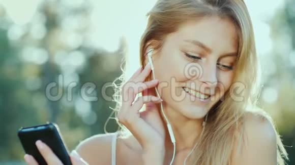 一位年轻的女士戴着耳机听音乐使用智能手机太阳美丽地照亮了她的头发视频的预览图