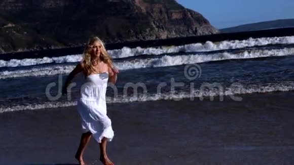 阳光明媚的一天美丽的金发女郎穿着白色连衣裙在海滩上奔跑视频的预览图
