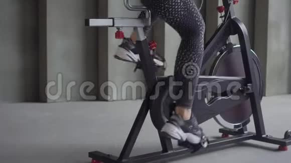 性感的年轻女子穿着黑色运动服在健身房骑自行车听着耳机里的音乐视频的预览图
