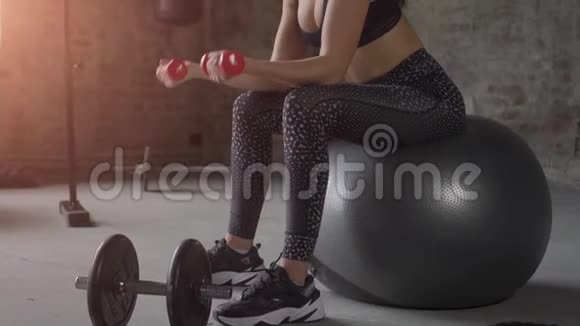 穿着黑色运动服的女孩在砖墙的背景下坐在瑞士球上用哑铃锻炼身体视频的预览图