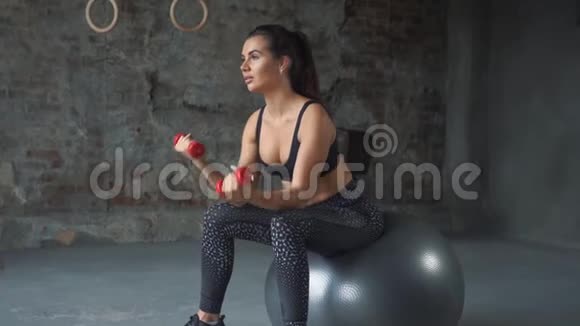 穿着黑色运动服的女孩在砖墙的背景下坐在瑞士球上用哑铃锻炼身体视频的预览图