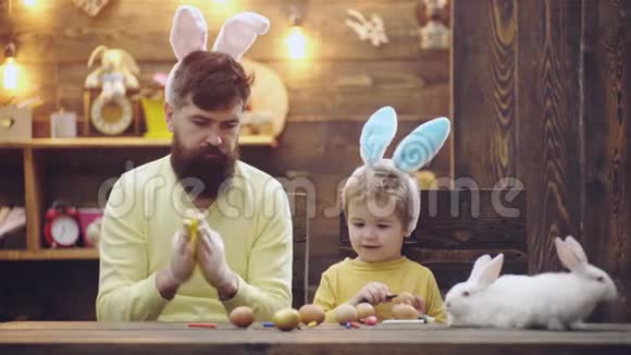 长胡子的男人和他的儿子穿着兔子耳朵装饰复活节彩蛋父子俩正在画蛋幸福的家庭视频的预览图
