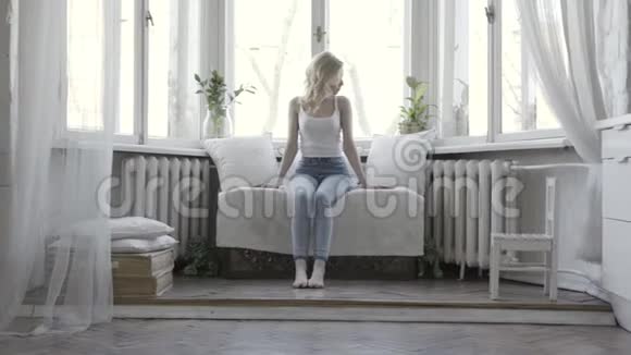 悲伤的年轻女子坐在沙发上在她明亮的公寓的大窗户前思考行动金发美女视频的预览图