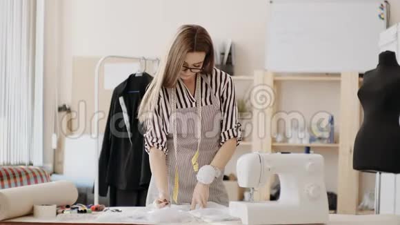女裁缝眼镜和围裙站在缝纫机附近的办公桌后面在缝纫机上做铅笔标记视频的预览图