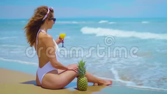 快乐美丽的小女孩坐在海边的沙滩上拿着菠萝和果汁视频的预览图