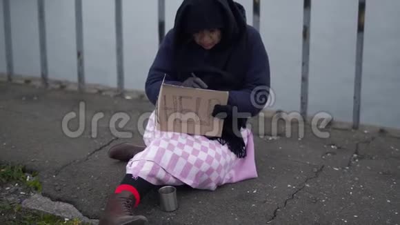 成年无家可归的妇女坐在桥上在寒冷多风的灰色天气里请求施舍和帮助并在桥上扭动一些东西视频的预览图