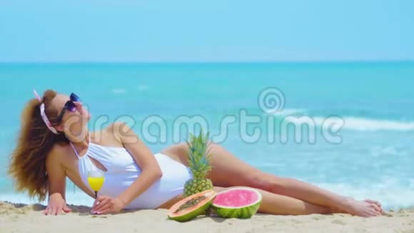 年轻漂亮的女人躺在沙滩上喝着橙汁概念假期视频的预览图