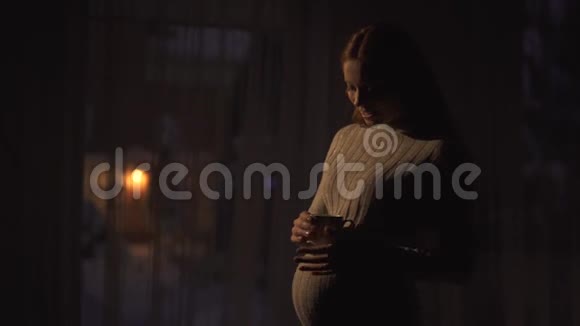一位留着金色长发的漂亮孕妇站在黑暗的房间里端着一杯茶视频的预览图