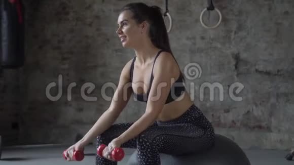 穿着黑色运动服的女士在砖墙的背景下坐在瑞士球上用哑铃锻炼身体视频的预览图