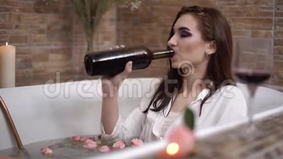 年轻悲伤的女人带着鲜艳的妆容喝着瓶子里的酒躺在花浴里视频的预览图