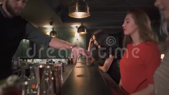 三个穿着漂亮的年轻女孩坐在酒吧旁边漂亮的时髦酒保一个接一个地给她们一杯鸡尾酒视频的预览图