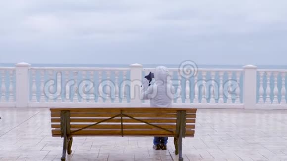 穿着白色夹克和引擎盖的女人在坐在美丽的带海的露台上的长凳上的智能手机上拍摄海浪视频视频的预览图