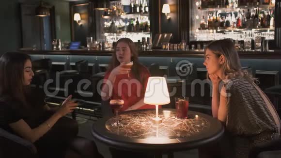三个穿着晚礼服的年轻女朋友坐在酒吧的小桌子旁女孩们一起庆祝视频的预览图