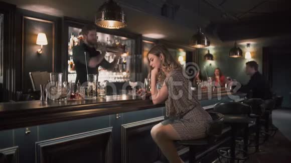 漂亮的女孩穿着闪亮的连衣裙坐在酒吧柜台前自拍在后台一个大胡子的酒保在发抖视频的预览图