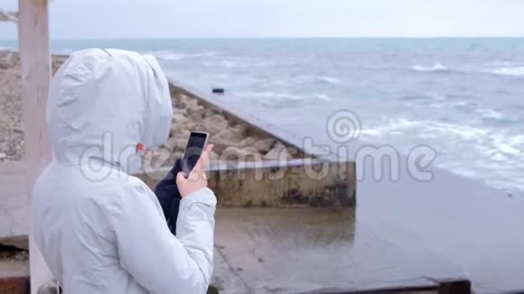 穿白色夹克的女人拿着手机大风浪站在堤岸上后景视频的预览图