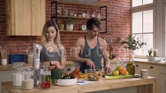 一对穿着运动服的年轻夫妇站在餐桌后面准备着新鲜的沙拉里面放着蔬菜视频的预览图