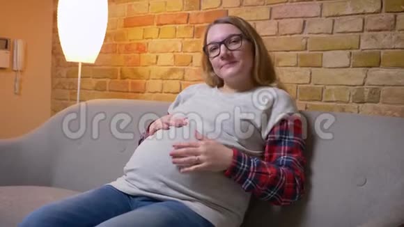 年轻怀孕女性坐在沙发上看镜头时轻轻抱着肚子的特写镜头视频的预览图