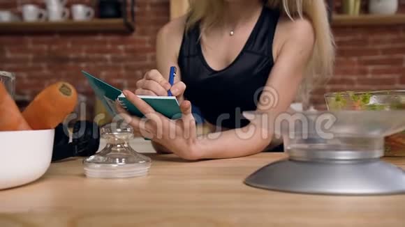 一位漂亮的年轻女士坐在厨房的桌子后面并在笔记本上记录食谱以促进健康视频的预览图