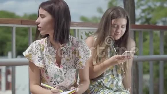 两个可爱的年轻女学生坐在户外一个女孩带着耳机听音乐第二个女孩视频的预览图