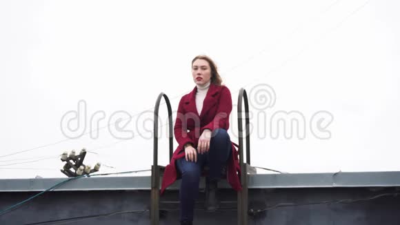 穿着红色外套白色高领毛衣和牛仔裤坐在屋顶楼梯上的年轻迷人女人的特写媒体照片视频的预览图