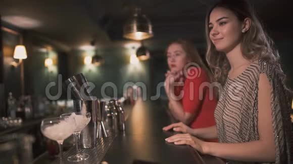 两个女朋友坐在酒吧柜台等酒保给他们做鸡尾酒美丽女孩的休闲视频的预览图